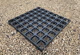 A square black plastic grid mat with circular cutouts, shown on a bed of small tan gravel stones.
