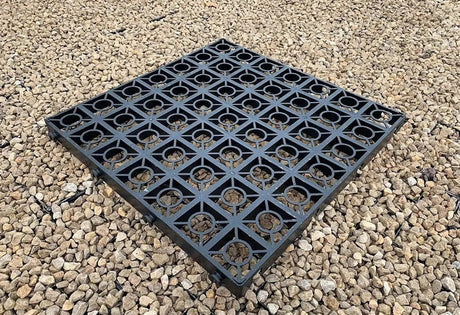 A square black plastic grid mat with circular cutouts, shown on a bed of small tan gravel stones.