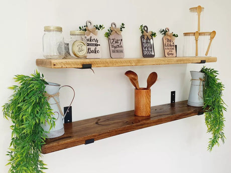 Two rustic wooden floating shelves mounted on a white wall, styled with decorative signs, glass jars, metal canisters, wooden utensils, and green trailing plants.
