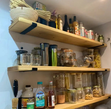 Three light wood floating shelves mounted on a white wall, styled with kitchen items including glass jars, bottles, a woven basket, and dried wheat stalks.